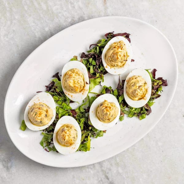 Six deviled eggs with paprika and herbs on a bed of mixed greens, served on a white oval plate.