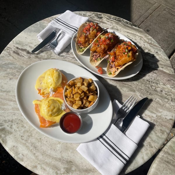 Two plates of American brunch food on a marble table, including eggs benedict and tacos.
