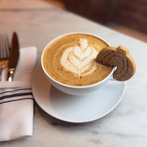 A latte with foam art and a pumpkin-shaped cookie on the rim, next to silverware.