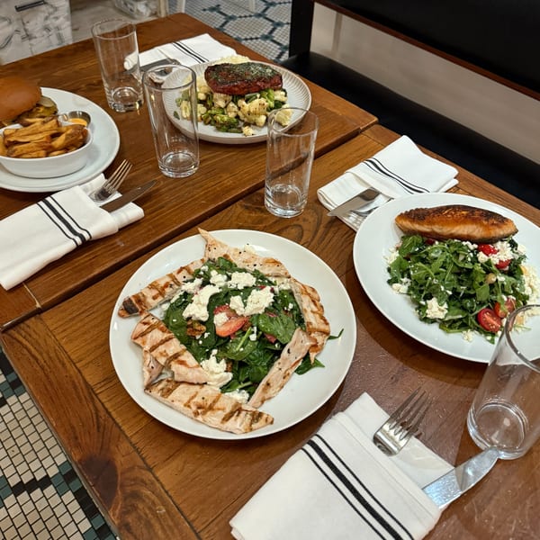Three American dishes on a wooden table: a burger with fries, grilled salmon salad, and chicken salad.