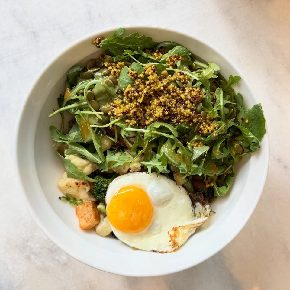 A White Maple Café breakfast bowl with arugula, quinoa, pumpkin seeds, roasted vegetables, and a fried egg.