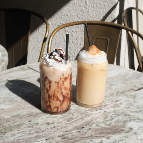 Two iced coffee drinks, one with whipped cream and chocolate, on a marble table outdoors.