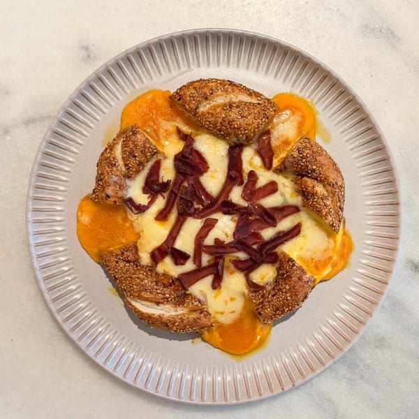 A savory pastry with sesame crust, melted cheese, cured meat, and a runny egg yolk on a plate.