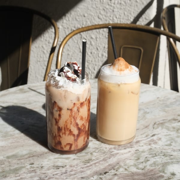 Two iced coffee drinks, one with whipped cream and chocolate drizzle, on a marble table.