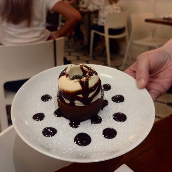 A hand holding a white plate with a chocolate lava cake topped with vanilla ice cream and chocolate syrup.