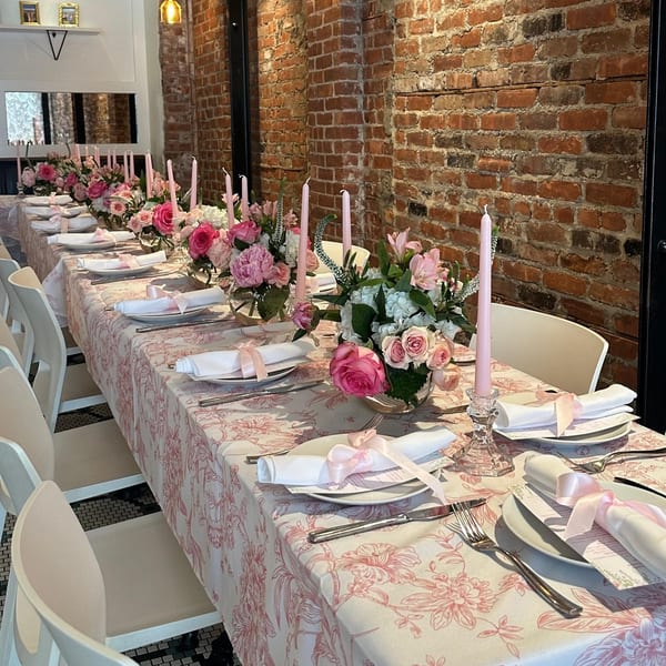 Long dining table set with pink floral tablecloth, candles, and flower arrangements at White Maple Café.