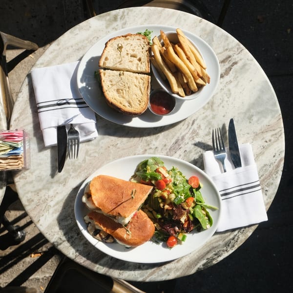Two American dishes, a sandwich with fries and a panini with salad, on a marble table.