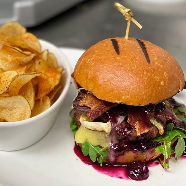 Gourmet burger with bacon, brie, arugula, and berry compote, served with a side of chips.
