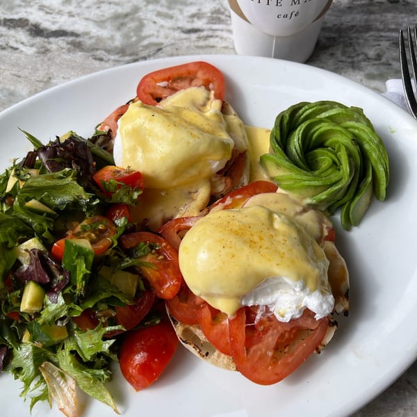 Eggs Benedict with sliced tomatoes, a side salad, and fanned avocado on a white plate.