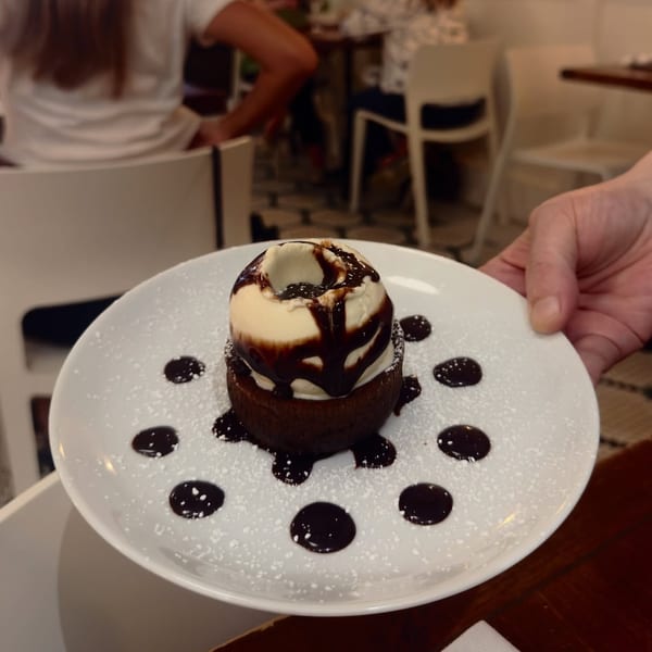 A hand holds a white plate with a chocolate lava cake topped with vanilla ice cream and chocolate syrup.