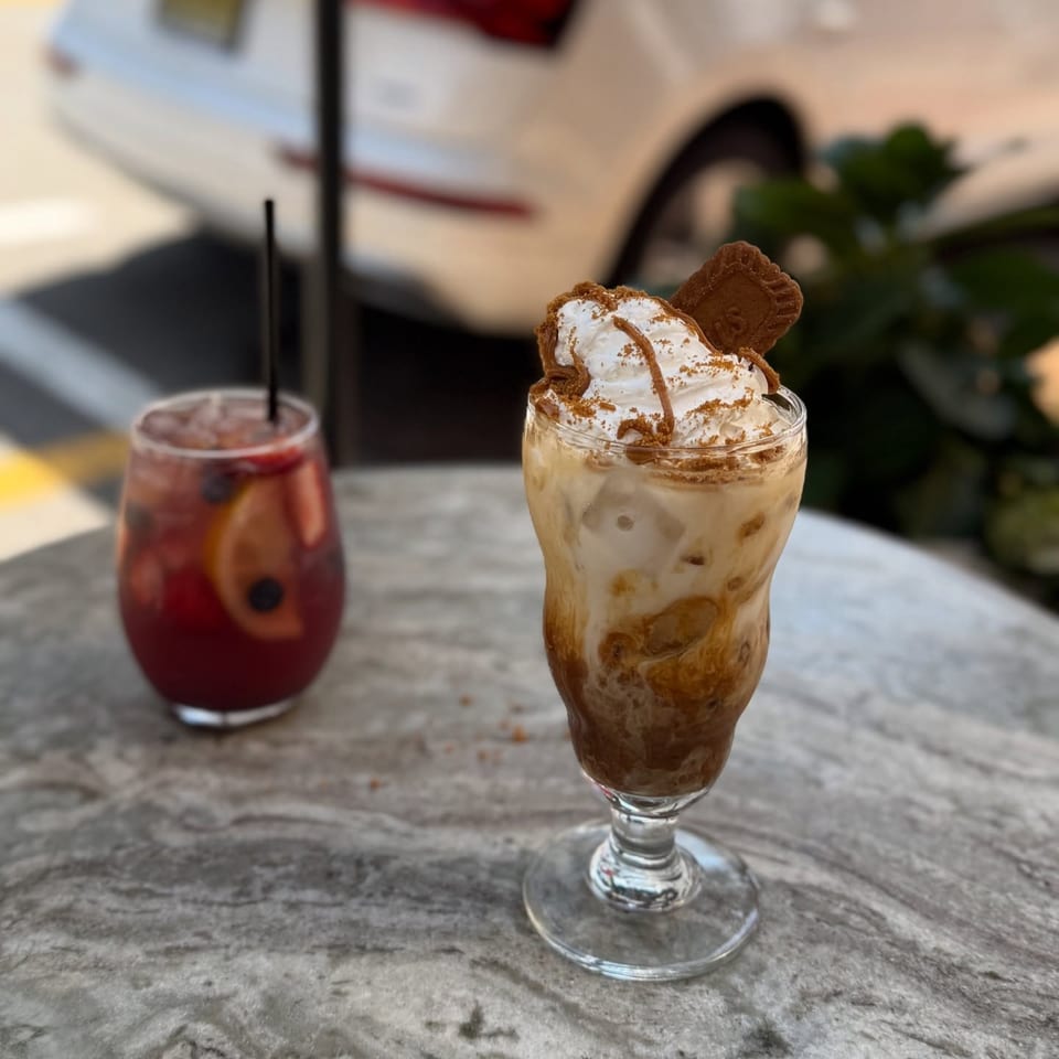 Two drinks on a marble table, one a red fruit drink and the other an iced coffee with whipped cream.